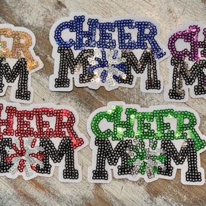 May include: Five sequined patches with the words "CHEER MOM" in various colors: gold, blue, purple, red, and green. The patches have a black border and a snowflake design in the center.