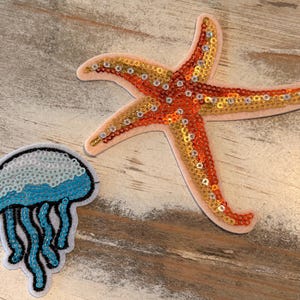 May include: Two sequined patches: a starfish and a jellyfish. The starfish is orange, gold, and pink. The jellyfish is white, blue, and black. Both patches are on a light brown, textured surface.