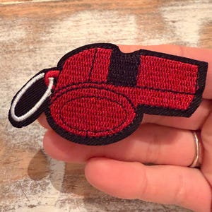May include: A red and black embroidered whistle patch with a black border and a white and black loop. The patch is in the shape of a whistle and is likely for a sports uniform or a craft project.