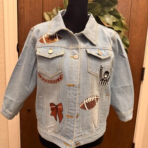 May include: A light blue denim jacket featuring various patches including two brown footballs, a red bow, a referee figure, and a patch with the text TOUCHDOWN. The jacket has a collar, long sleeves, two chest pockets with flaps, and silver buttons.