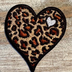 May include: Embroidered heart-shaped patch with a leopard print pattern in shades of brown and tan, outlined in black. A small white heart is embroidered on the right side. This patch is suitable for clothing and accessories.