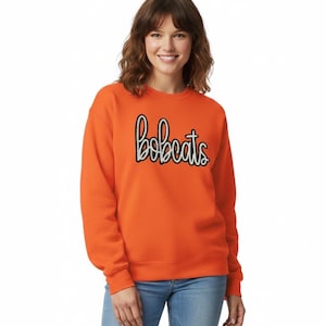 May include: An orange sweatshirt with the word "bobcats" in a script font. The text is outlined in black and white. The sweatshirt is a solid orange color and is being worn with blue jeans.