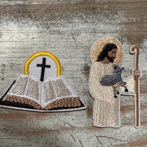 May include: Two embroidered patches, one depicting a white open book with a gold halo and a black cross, and the other depicting a white figure with a brown beard holding a lamb and a brown staff.