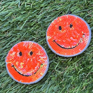 May include: Two orange smiley face patches with sequins. The patches are on a green artificial grass background.