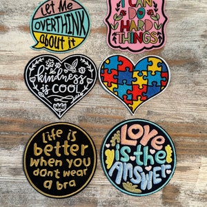 May include: Six embroidered iron-on patches with various sayings. The patches are in different shapes and colors, including a heart, a speech bubble, and a puzzle piece. The sayings include "Let me overthink about it", "I can do hard things", "Kindness is cool", "Life is better when you don't wear a bra", "Love is the answer", and "I can do hard things".