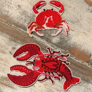 May include: Two embroidered patches featuring sea creatures. The top patch is a red crab with white and pink accents. The bottom patch is a red lobster with intricate detailing. Both patches are on a light-colored, textured surface.