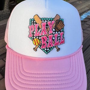 May include: A pink and white trucker hat with a white mesh back. The front of the hat features an embroidered patch with a baseball, bats, and the words "PLAY BALL" in pink letters.