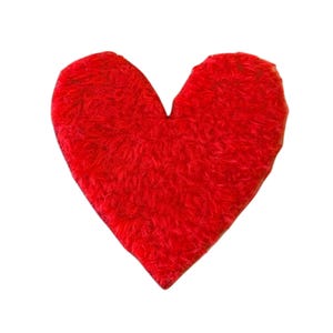 May include: A heart-shaped patch or applique featuring a vibrant red color and a dense, fuzzy, textured surface. The object has a distinct, symmetrical heart silhouette with a rounded top and a pointed bottom, showcasing a soft, plush appearance.