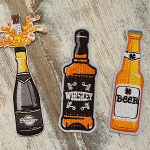 May include: Embroidered patches featuring a champagne bottle with a golden spray, a whiskey bottle with the word "Whiskey", and a beer bottle with the word "Beer". The patches have a white border and are on a wooden surface.