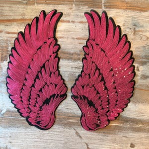 May include: A pair of mirror-image wing-shaped patches covered in vibrant hot pink sequins. Each patch features a detailed feather pattern with a black fabric border outlining the shape, creating a distinct contrast against the bright pink surface.