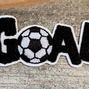 May include: A black and white embroidered patch with the word "GOAL" in bold, block letters. The letter "O" is replaced by a soccer ball design. The patch has a white border and is set against a textured, light brown surface.