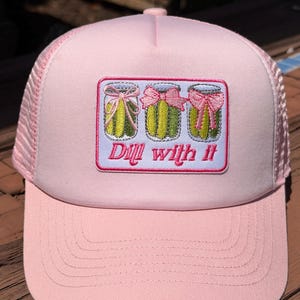 May include: A light pink trucker hat featuring an embroidered rectangular patch. The patch displays three glass jars containing green pickles, each adorned with a pink ribbon bow. Below the jars, the text reads Dill with it in pink script.