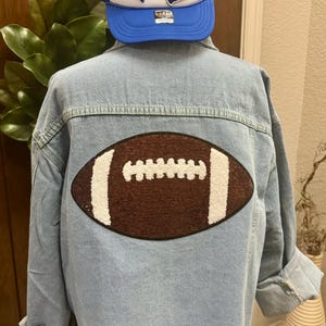 May include: A light blue denim jacket with a brown and white football patch on the back. The jacket has a frayed hem and a blue and white trucker hat with a heart, football, and megaphone patch is on top of the jacket.
