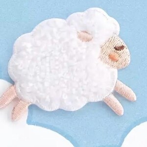 May include: An embroidered patch featuring a white sheep with a fluffy, textured wool body, light pink legs, and a light pink face with closed eyes and rosy cheeks. The sheep is depicted in a horizontal position against a light blue background.