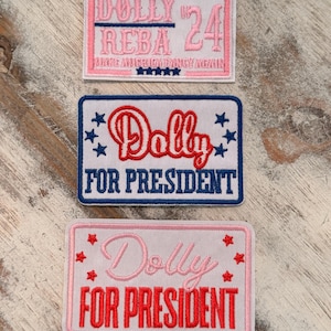 May include: Three rectangular embroidered patches. The top patch is pink with the text "DOLLY REBA 24" and "MAKE AMERICA DINGY AGAIN". The middle patch is white with "Dolly FOR PRESIDENT" in red and blue. The bottom patch is pink with "Dolly FOR PRESIDENT" in red.