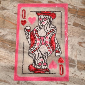 May include: A playing card featuring the Queen of Hearts, adorned with red and pink sequins. The card has a pink border and includes heart symbols and the letter "Q". The queen is depicted with heart-shaped eyes and a red hat.