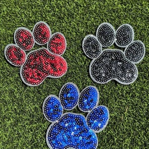 May include: Three sequined paw print patches in red, black, and blue. Each paw print has a silver glitter border. The patches are on a green, grassy surface.