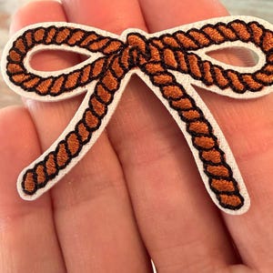 May include: An embroidered patch in the shape of a bow, featuring a copper-colored rope design with black outlines on a white fabric backing. The bow has two loops and two trailing ends, with the rope texture clearly defined by stitching.