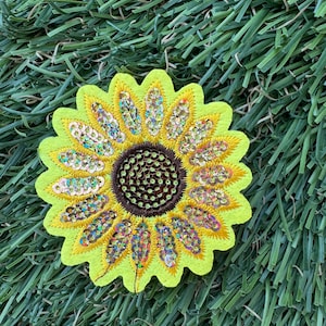 May include: A yellow felt sunflower with a brown center and iridescent sequins. The sunflower is on a green artificial grass background.