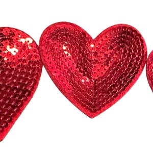 May include: Three red sequined heart-shaped patches arranged in a row. The hearts vary slightly in size, with the largest on the left and the smallest on the right. The sequins catch the light, creating a shimmering effect against the white background.