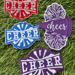 May include: Four iron-on patches with a cheer theme. The patches are red, blue, purple, and white. The patches feature the word "cheer" in white lettering. One patch features a purple megaphone with a white pom-pom.
