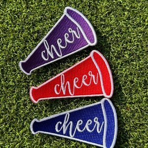 May include: Three embroidered iron-on patches shaped like megaphones with the word "cheer" written in white on each. The megaphones are purple, red, and blue.