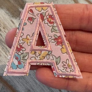 May include: A pink fabric letter "A" patch with a floral pattern in red, yellow, and blue. The letter is outlined with a pink border, creating a decorative and personalized element.