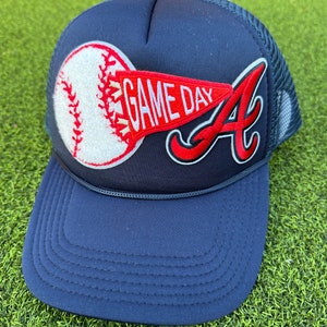 May include: A navy blue trucker hat with a white baseball and red "GAME DAY" pennant patch. The hat also features a red "A" patch.