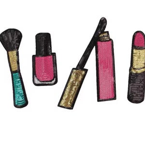 May include: A collection of sequined makeup patches, including a black makeup brush with a gold handle and teal base, a pink nail polish bottle, a pink lip gloss, and a pink lipstick with a gold base.