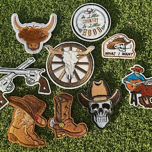 May include: A set of nine embroidered iron-on patches featuring a variety of western and country themes. The patches include a cowboy boot, a skull wearing a cowboy hat, a skull with antlers, a highland cow, a cowboy playing a guitar, two pistols, and two patches with text that reads "A Little Country A Little Hood" and "I Do A Thing Called What I Want".