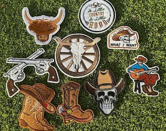 May include: A set of nine embroidered iron-on patches featuring a variety of western and country themes. The patches include a cowboy boot, a skull wearing a cowboy hat, a skull with antlers, a highland cow, a cowboy playing a guitar, two pistols, and two patches with text that reads "A Little Country A Little Hood" and "I Do A Thing Called What I Want".