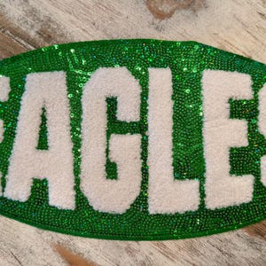 May include: An oval-shaped patch with the word "EAGLES" in white, fluffy letters against a background of green sequins. The patch is likely for a sports team or school.