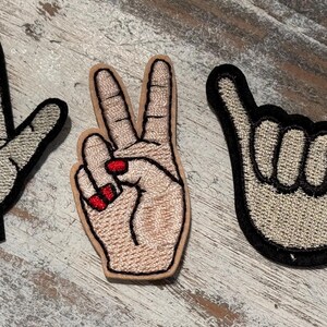 May include: Three embroidered iron-on patches featuring hand gestures. The patches are beige with black outlines. One patch shows a hand giving the 'rock on' sign, another shows a hand giving the 'peace' sign, and the third shows a hand giving the 'hang loose' sign.