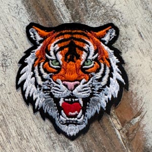 May include: Embroidered tiger patch with orange, black, white, and green details. The tiger's face is centered, with an open mouth revealing a red interior. The patch has a black border and is likely for clothing or accessories.