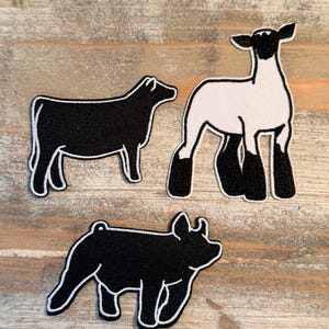 May include: Three fabric patches featuring farm animals: a black cow, a white and black lamb, and a black pig. Each patch has a white outline, creating a contrasting border. The patches are set against a light brown, wood-grain background.