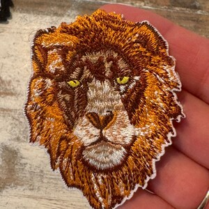 May include: Embroidered lion head patch with a brown and orange mane and yellow eyes. The patch is on a white background.