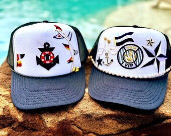 May include: Two trucker hats with white foam fronts and dark gray brims. The left hat features a black anchor and red life preserver patch. The right hat displays a circular VIP patch, a nautical star, and a string of white faux pearls along the brim.