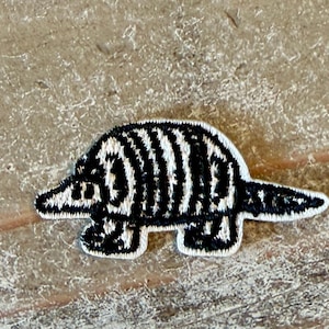 May include: A small embroidered patch featuring a stylized armadillo in black and white thread. The design shows the animal in profile with a pointed snout, a rounded shell with vertical stripes, four short legs, and a tapered tail, all outlined in white.