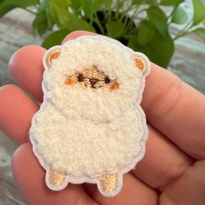 May include: A white, fluffy sheep-shaped embroidered patch with gold accents on the ears and feet. The sheep has a brown face and is held in a hand against a blurred green background.