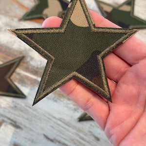 May include: A star-shaped fabric patch featuring a camouflage pattern in shades of olive green, dark brown, and tan. The patch has a dark green embroidered border along all five edges, providing a finished look to the star-shaped textile design.