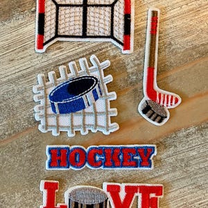 May include: A collection of embroidered patches featuring a red and white hockey goal, a blue hockey puck on a net, a hockey stick with a puck, and two text patches reading HOCKEY and LOVE, with the letter O in LOVE replaced by a hockey puck.