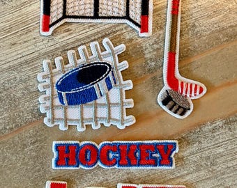 May include: A collection of embroidered patches featuring a red and white hockey goal, a blue hockey puck on a net, a hockey stick with a puck, and two text patches reading HOCKEY and LOVE, with the letter O in LOVE replaced by a hockey puck.