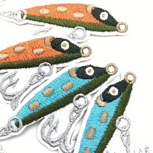 May include: Embroidered patches in the shape of colorful fish lures. The patches feature orange, blue, and green bodies with gold and black accents. Each patch has a silver embroidered hook.