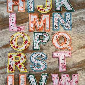 May include: A collection of fabric letters from A to Z, each with a floral pattern in various colors. The letters are outlined in different colors, including blue, orange, green, yellow, and pink, and are arranged on a wooden surface.