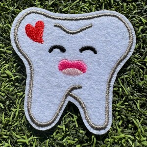 May include: A white felt tooth with a gray outline, smiling with closed eyes and a pink mouth. A red heart is stitched on the top left corner of the tooth.