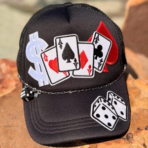 May include: A black trucker hat featuring embroidered patches of three playing cards, a light blue dollar sign, and two white dice. A silver-toned metal chain is attached across the front, adorned with a small black die charm on the left side.