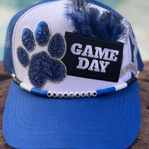 May include: A blue and white trucker hat with a blue sequined paw print, a black patch that says "GAME DAY", and a white beaded band that says "BOBOCATS".