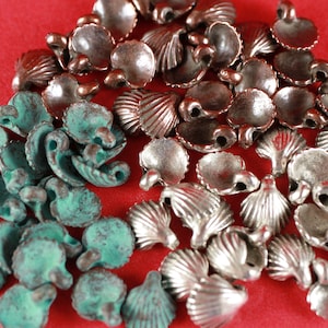 May include: A collection of metal shell-shaped charms in various shades of green, silver, and copper. The charms have a loop at the top for attaching to jewelry or other crafts.