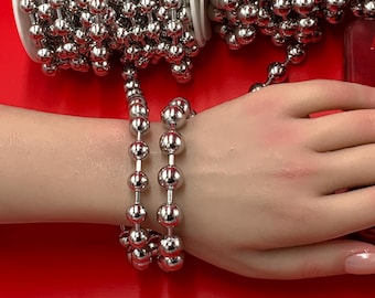 May include: Silver ball chain jewelry, displayed on a red surface. The chain is wound on white spools and also worn as a bracelet. The shiny, metallic beads are evenly spaced, creating a decorative and eye-catching design.