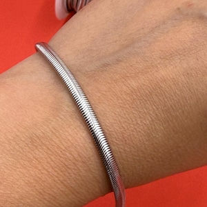 May include: A silver-colored metallic snake chain bracelet with a smooth, flexible, and segmented texture. The bracelet is shown alongside a white spool containing additional matching silver-colored metallic snake chain material.